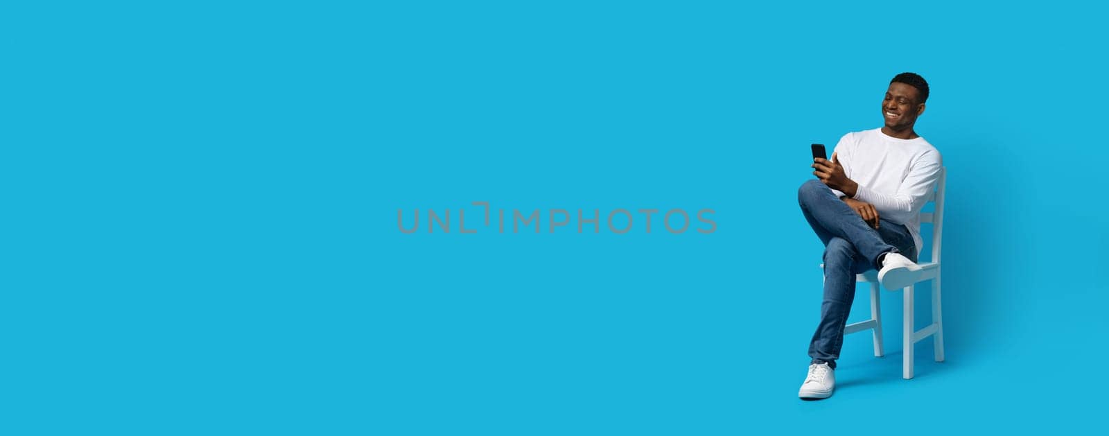A young African American man relaxes on a white chair while using his smartphone, showcasing a joyful expression. The bright blue backdrop enhances the vibrant mood of the moment.