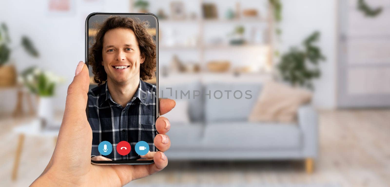 A hand holds a smartphone displaying a man smiling during a video call. The setting features a cozy home interior with a modern couch and plants in the background, creating a relaxed atmosphere.