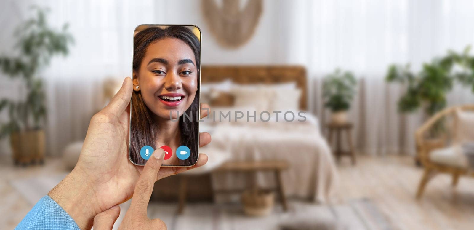 A person is holding a smartphone while having a video call in a cozy room. The background features a comfortable bed and plants, creating a warm atmosphere for conversation.