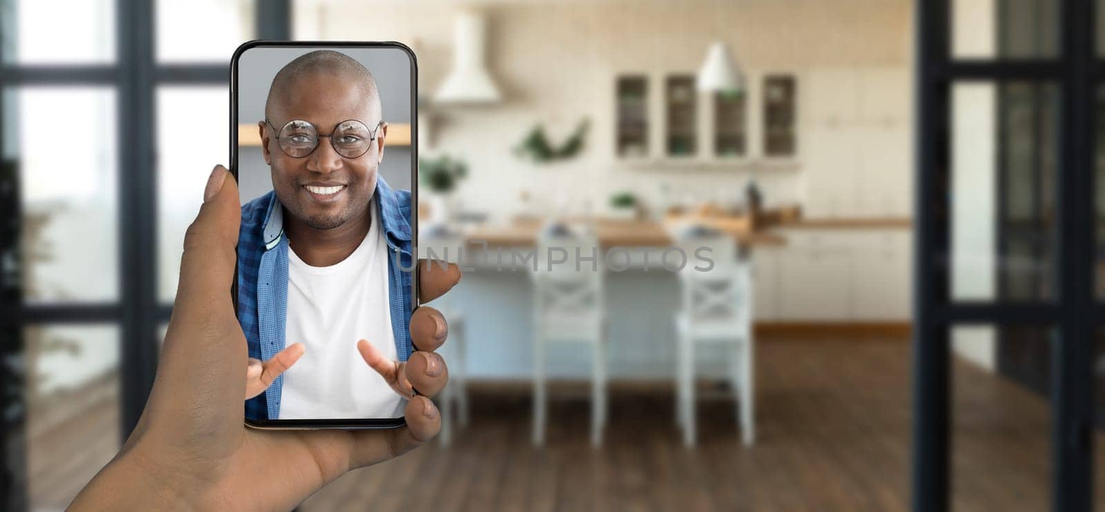 A person holds a smartphone displaying a smiling individual during a video call. The setting features a bright kitchen with wooden floors and a modern, inviting atmosphere.