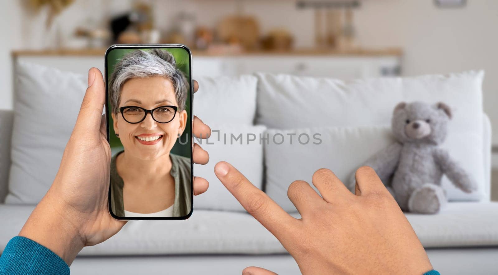 A person is using a smartphone for a video call from their home. The lady on screen smiling and engaged in conversation while relaxing on a couch with a teddy bear nearby.