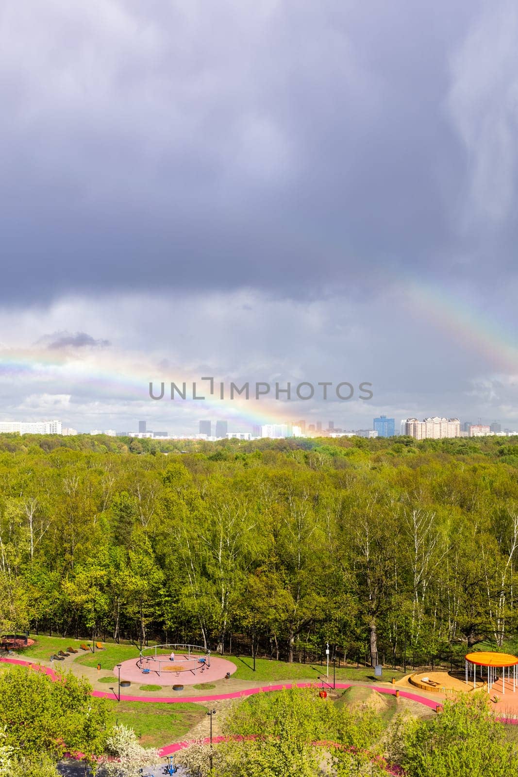 gray rainy clouds and two rainbow over sunlit playground near city park after rain on sunny spring day