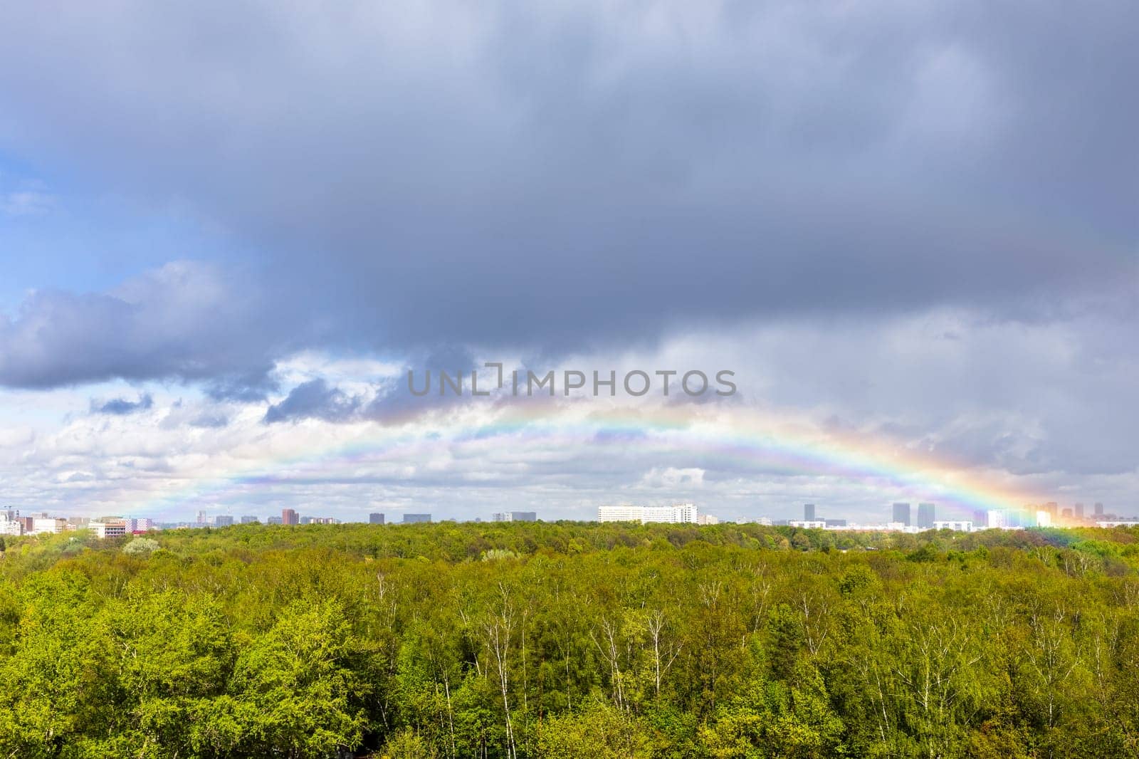 gray rainy clouds and rainbow in blue sky over green city park after rain on sunny spring day