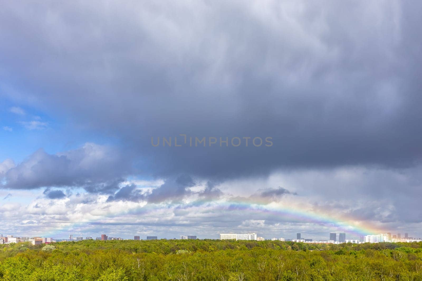 grey rainy clouds and rainbow over green city park after rain on sunny spring day