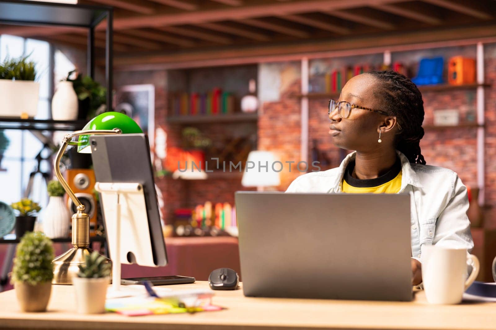 Contemplative woman daydreaming, gazing outside the window, feeling bored after working too much on job tasks. African american person at home caught in thoughts, pondering