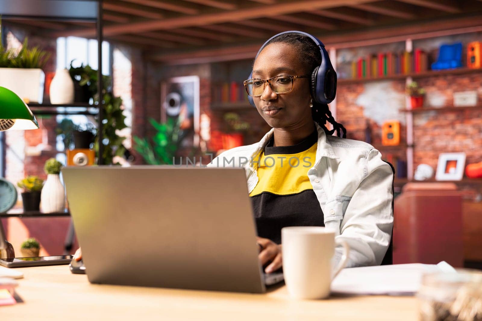 Person enjoying music while researching data for company project, doing tasks in personal home office. African american woman sitting at desk in front of laptop and using headphones