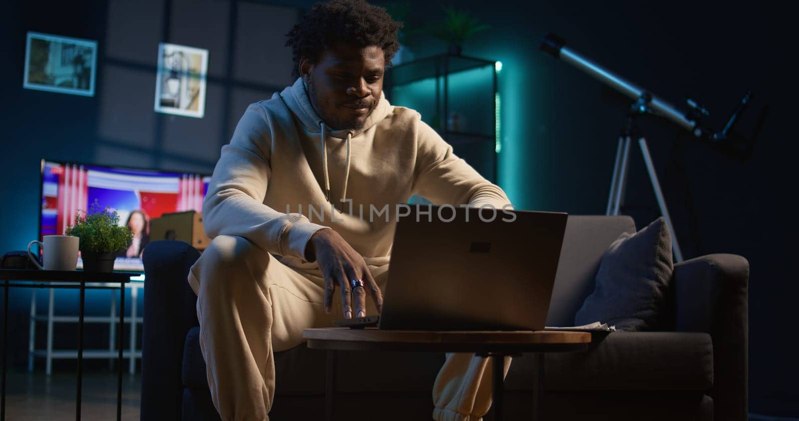 Ambitious black man learning a new language alone in his cozy living room, browsing internet for study platforms and explanations. Adult using language learning software online on laptop. Camera B.