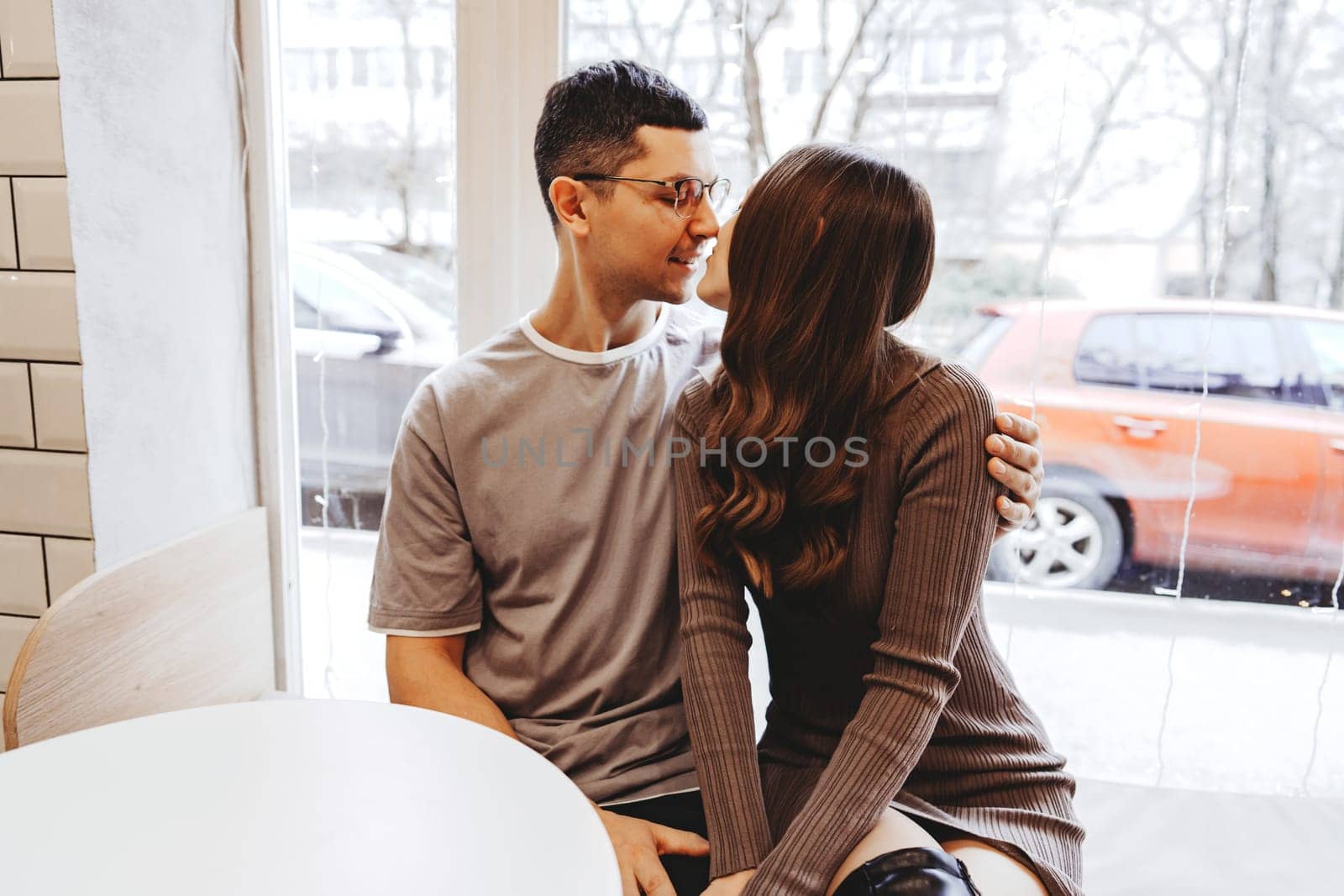 A couple shares a warm moment in a cafe, embracing and smiling at each other. Outside, a wintry scene unfolds with cars parked and snow falling gently.