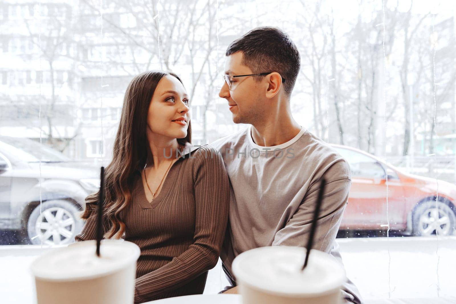Two individuals share a warm moment in a cafe, gazing into each other's eyes over coffee. Outside, snowflakes fall softly, creating a picturesque winter backdrop.
