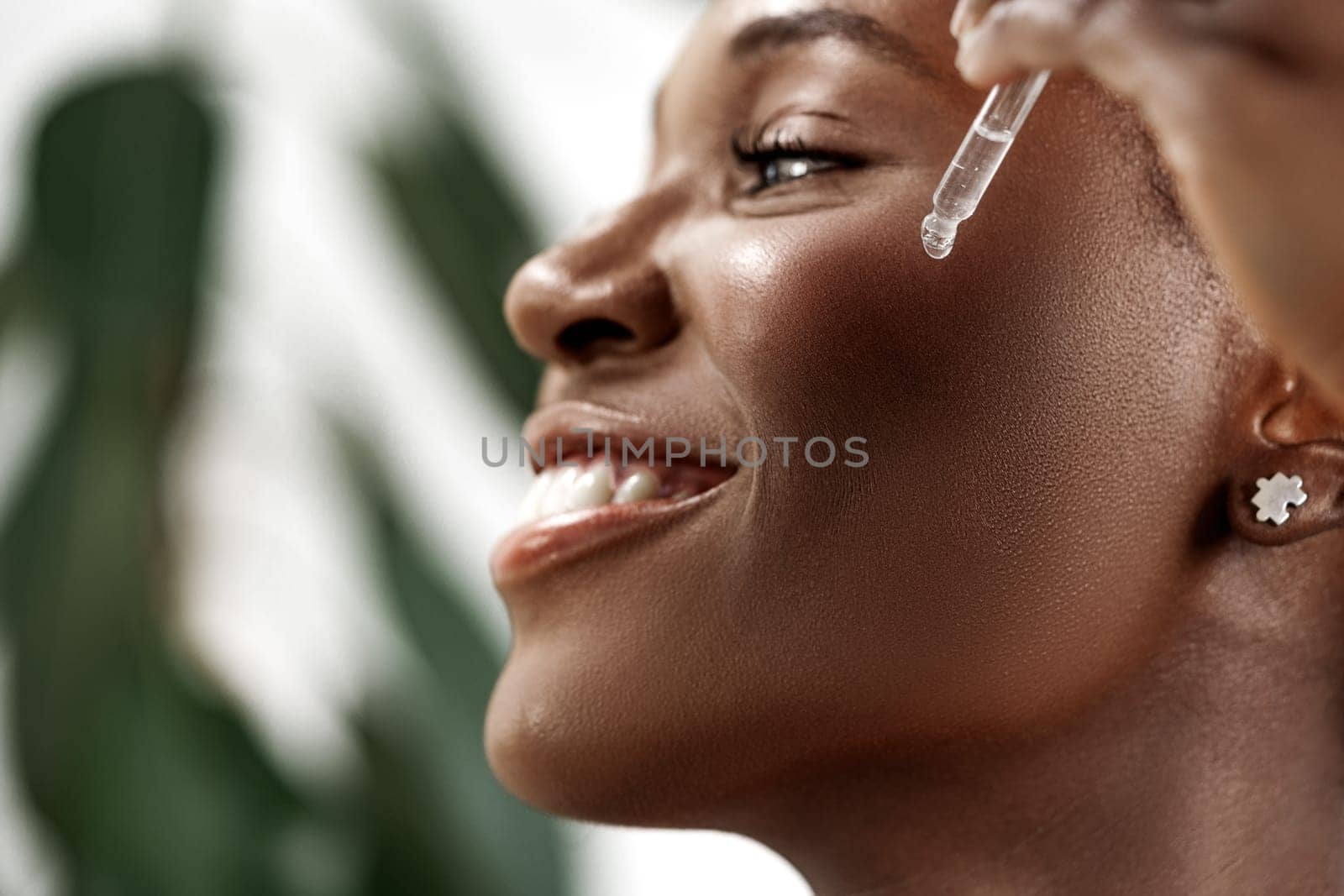 A woman with a joyful expression is applying a dropper serum to her face, surrounded by greenery. Sunlight highlights her radiant skin as she enjoys her skincare routine.