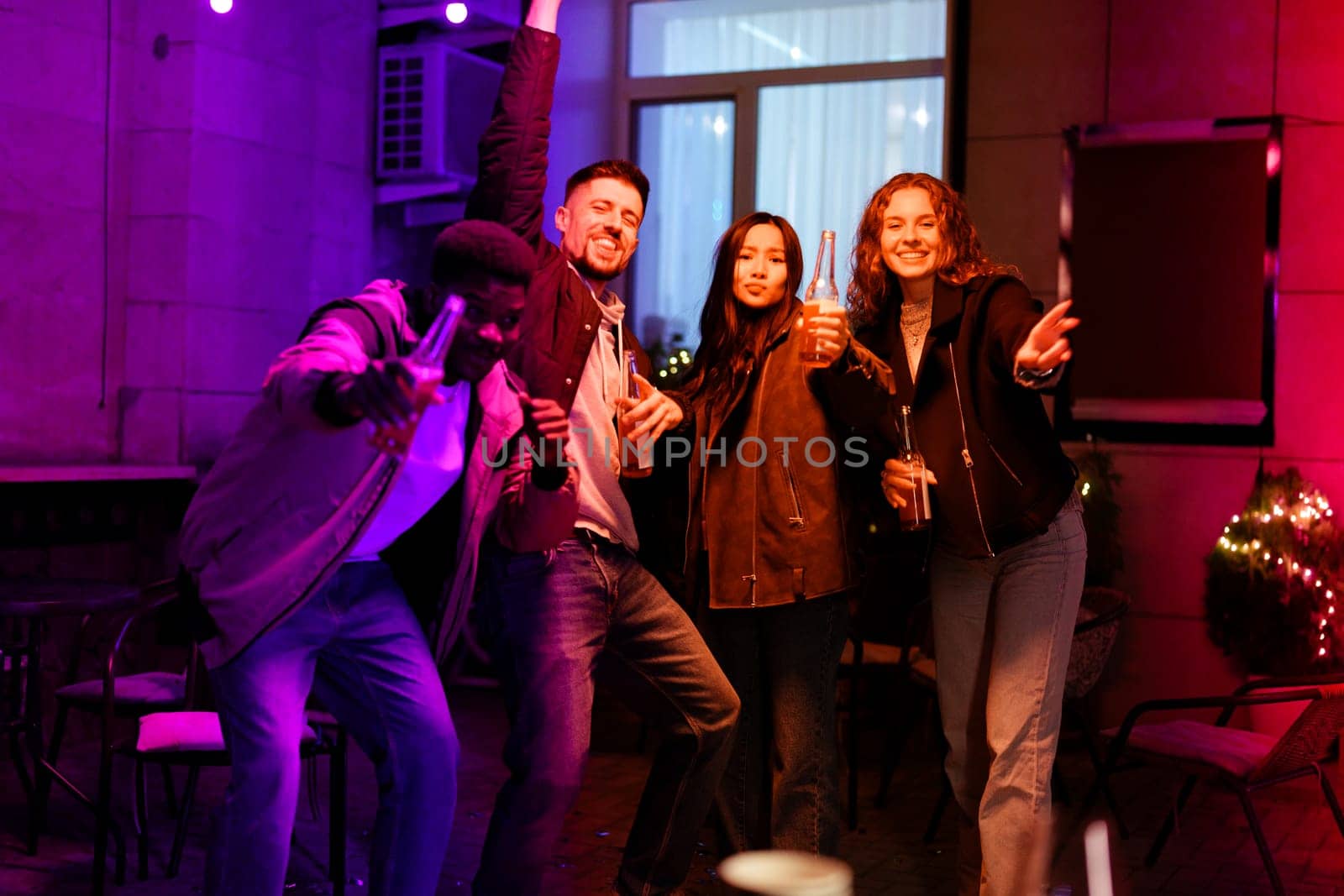 Four friends dance and celebrate together in a lively atmosphere filled with colorful lights. They enjoy drinks while showcasing their excitement and camaraderie at night.