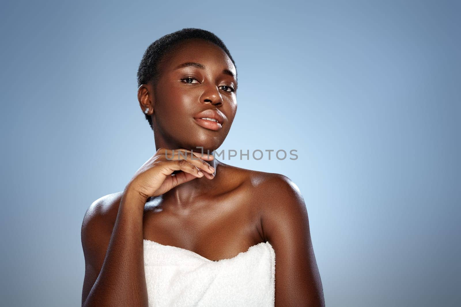 A woman stands gracefully with a soft expression, her short hair styled neatly. Draped in a white towel, she highlights her radiant skin and exudes confidence in a serene atmosphere.