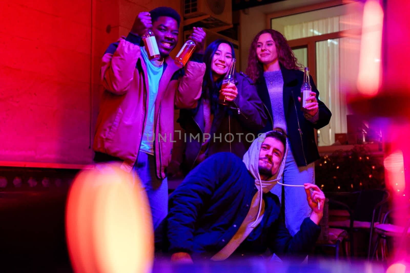 Four friends gather indoors, holding drinks while enjoying a vibrant atmosphere filled with colorful lights. They share smiles, embodying a joyous celebration together.