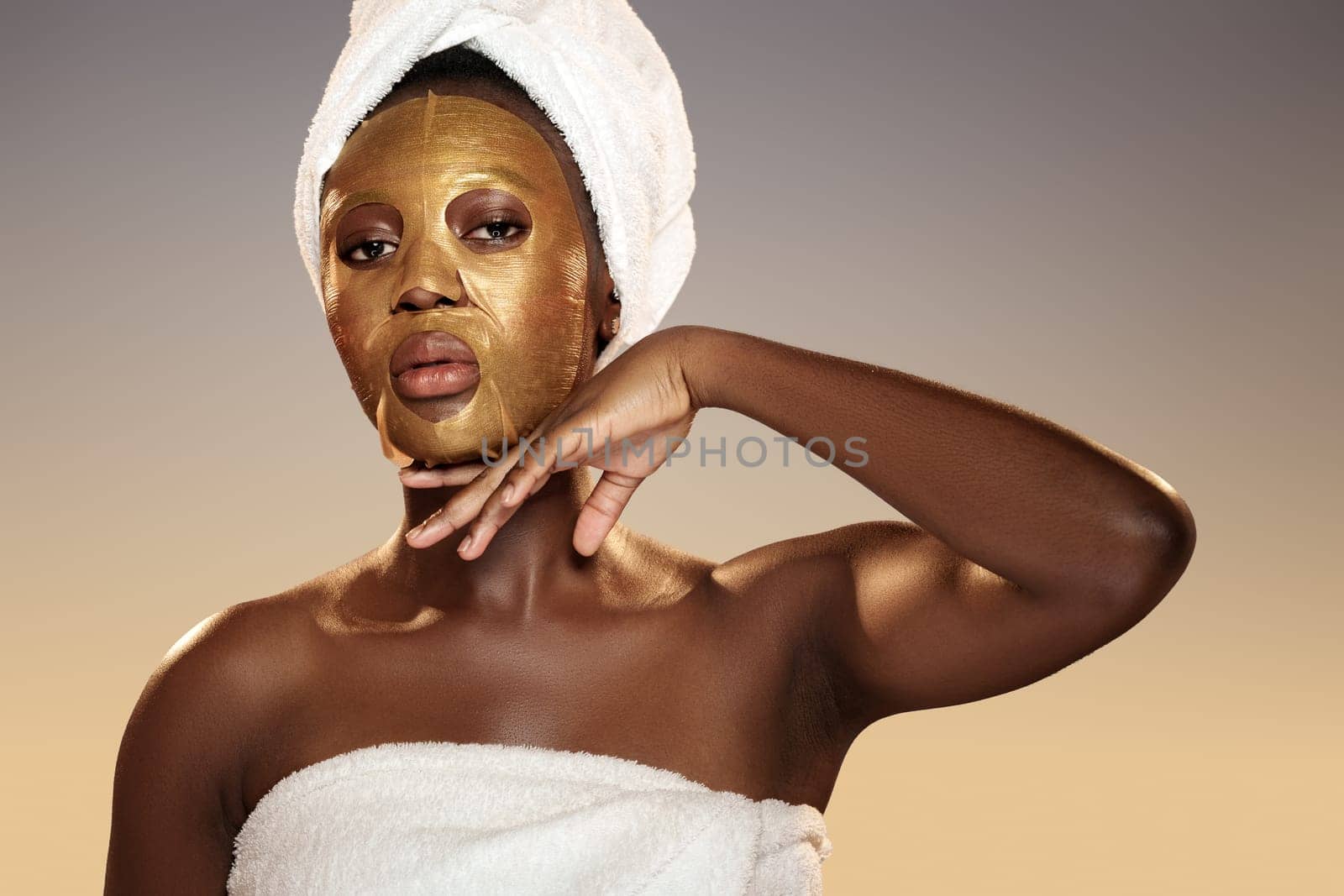 A woman enjoying self-care with a shiny gold face mask on her skin poses gracefully with her hand against her neck, surrounded by a calming atmosphere and soft lighting.