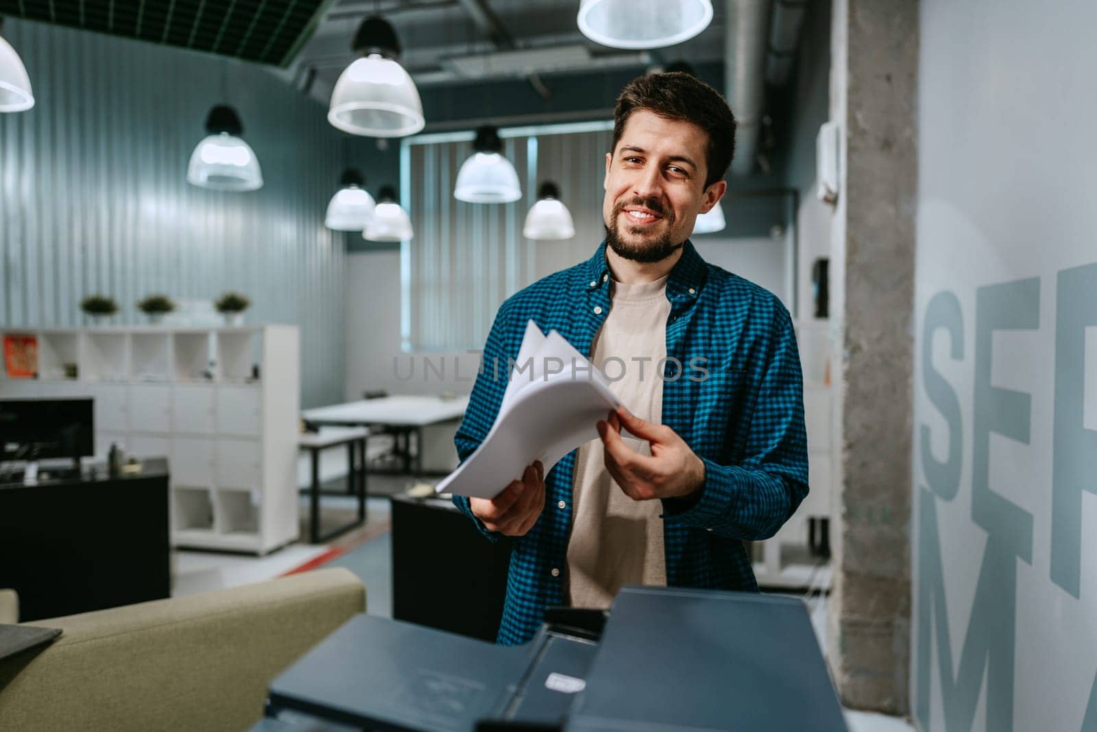 A man stands in a contemporary office environment, cheerfully sorting through papers. He wears a blue checkered shirt, surrounded by modern furnishings and warm lighting.