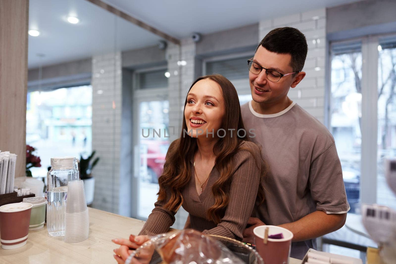 A couple shares a joyful interaction at a cafe counter, with bright windows letting in daylight. The cozy atmosphere enhances their connection and happiness.