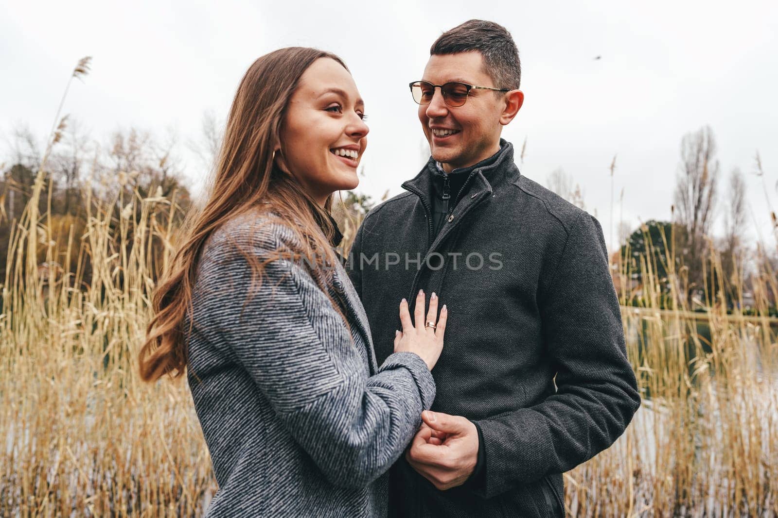 A couple stands by a serene lake, smiling and holding hands. Tall grass sways gently in the breeze, adding a peaceful ambiance to their affectionate interaction on a cloudy day.