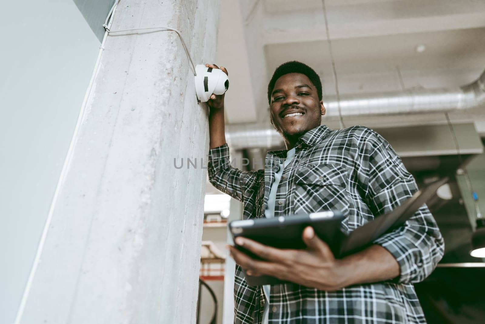A cheerful young man stands next to a concrete pillar, holding a tablet in one hand and gesturing towards a wall light. The contemporary workspace has a vibrant atmosphere.