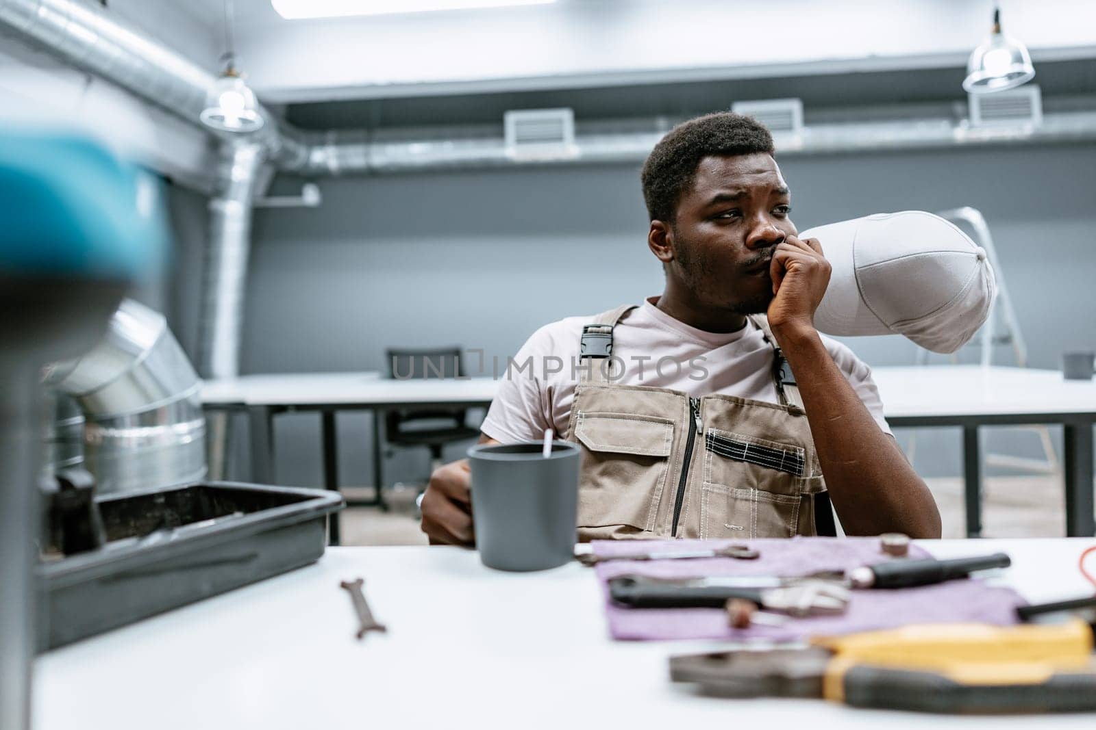 A man in work overalls sits at a table in a modern workshop. He holds a cup in one hand and looks thoughtful, surrounded by tools and equipment, during a brief pause.