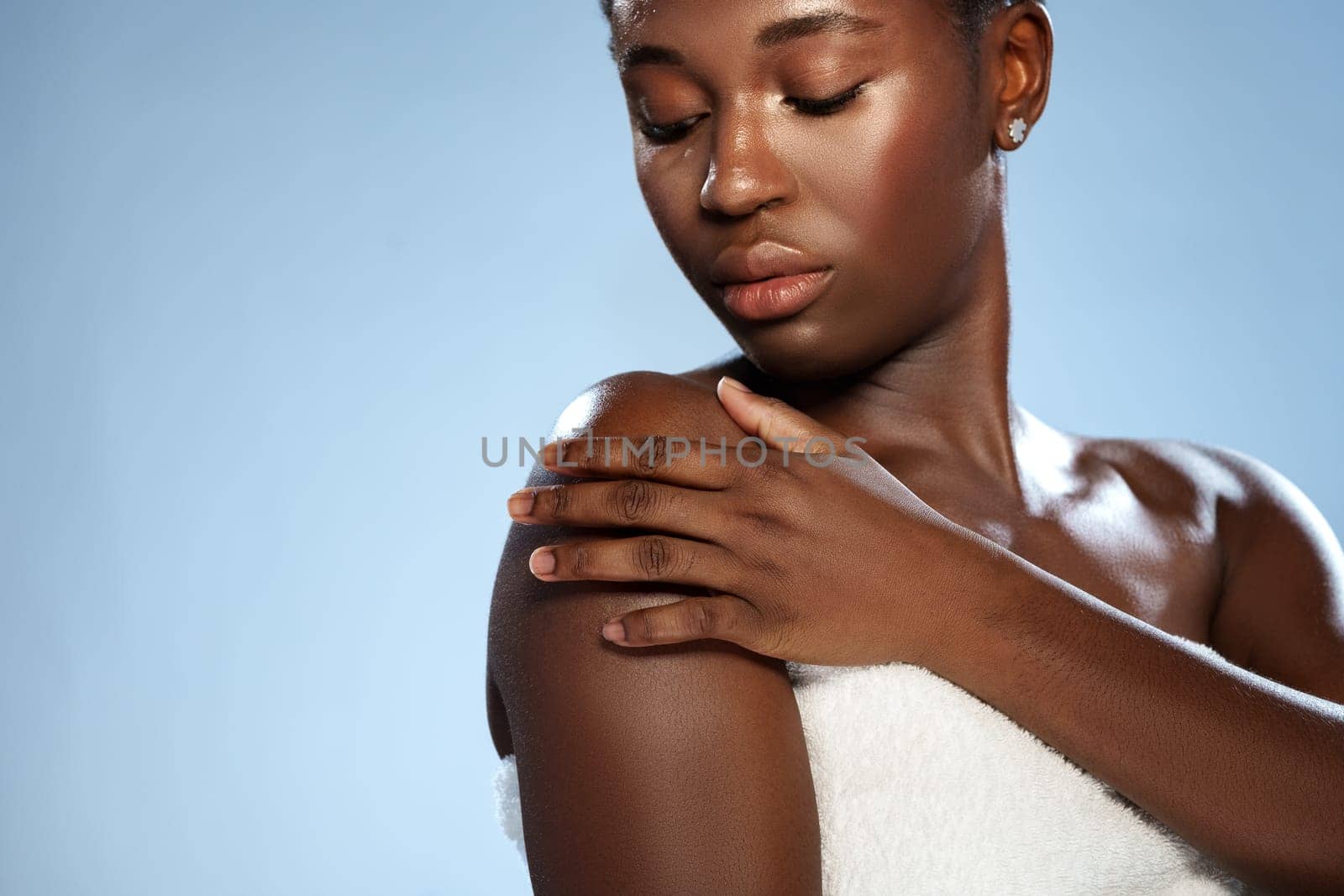 A young woman with smooth skin touches her shoulder delicately while posing. She has a serene expression and the soft light accentuates her natural beauty against a light blue backdrop.