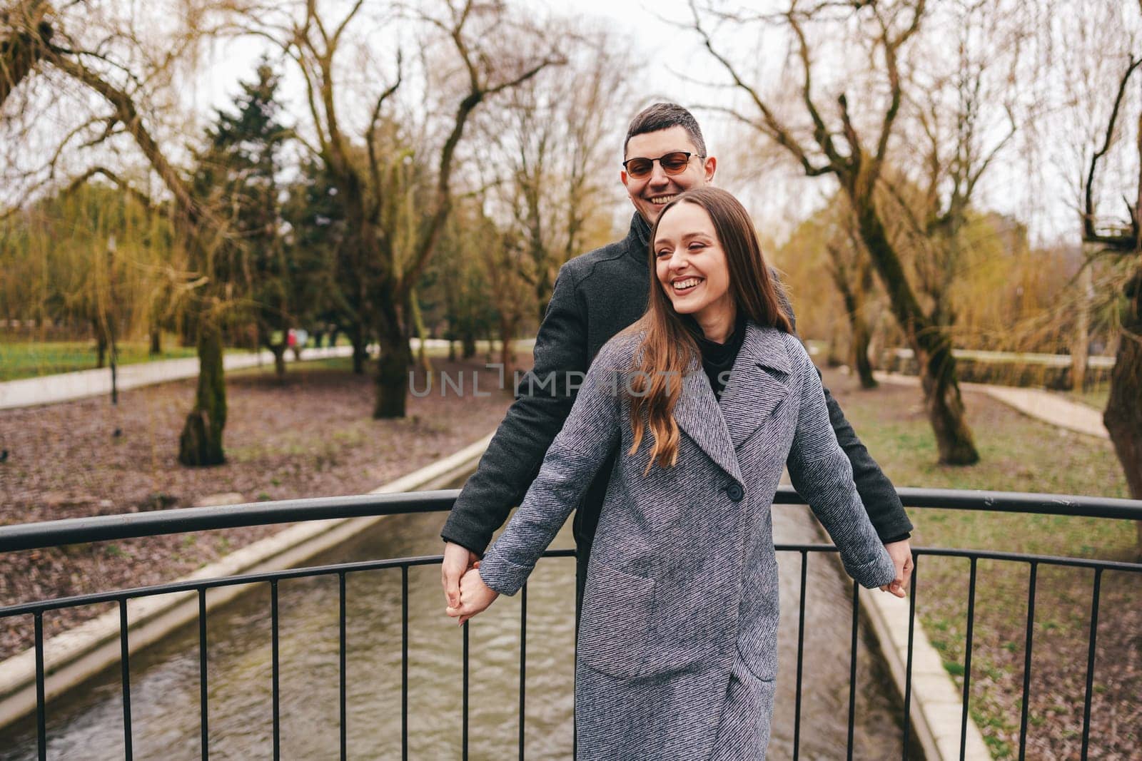 A couple happily holds hands on a bridge while smiling at each other, enjoying a serene day in a park with bare trees and a gentle stream flowing below.