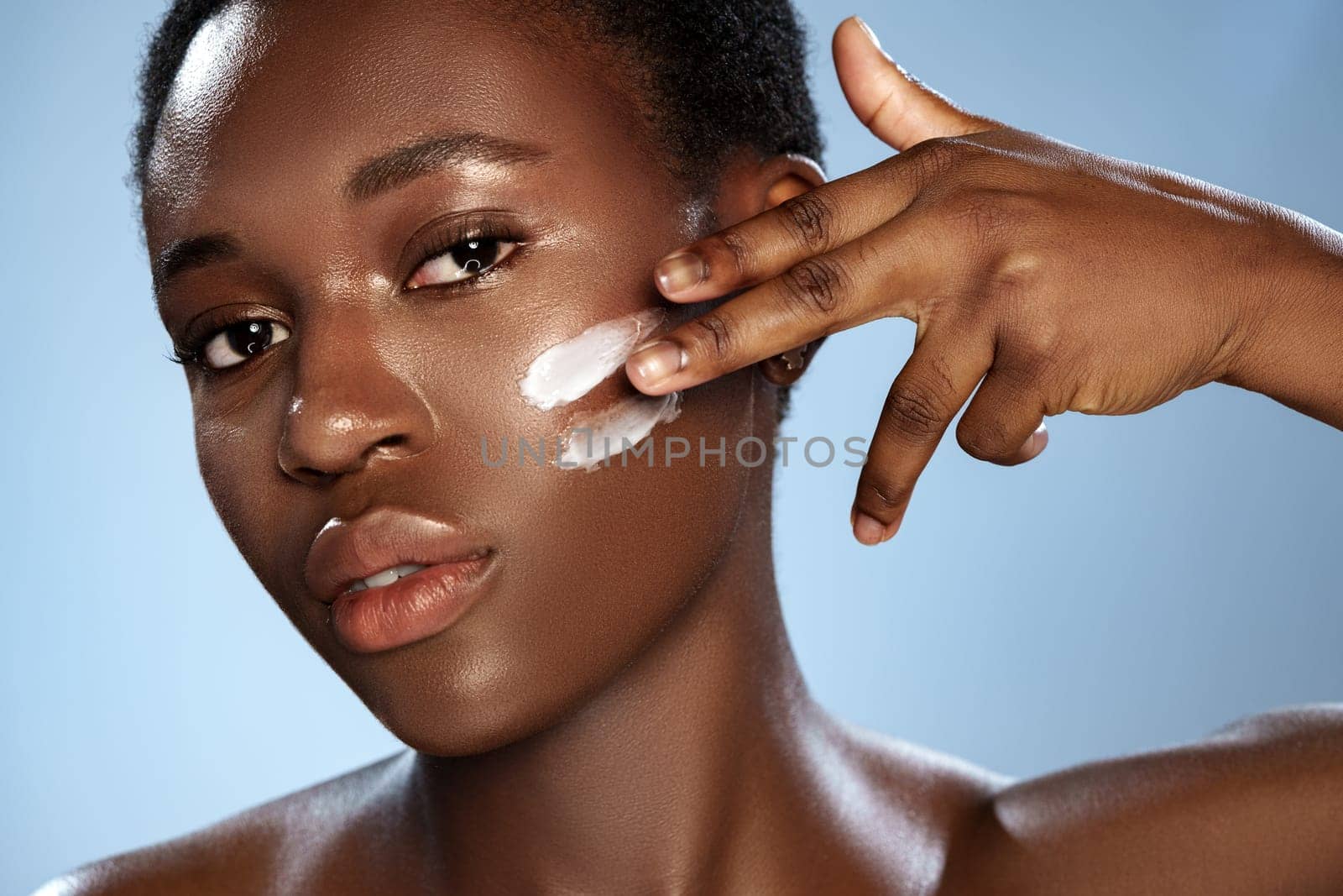 A woman with short natural hair applies a creamy skincare product to her cheek in a bright studio. The soft blue backdrop enhances her radiant complexion and focused expression.