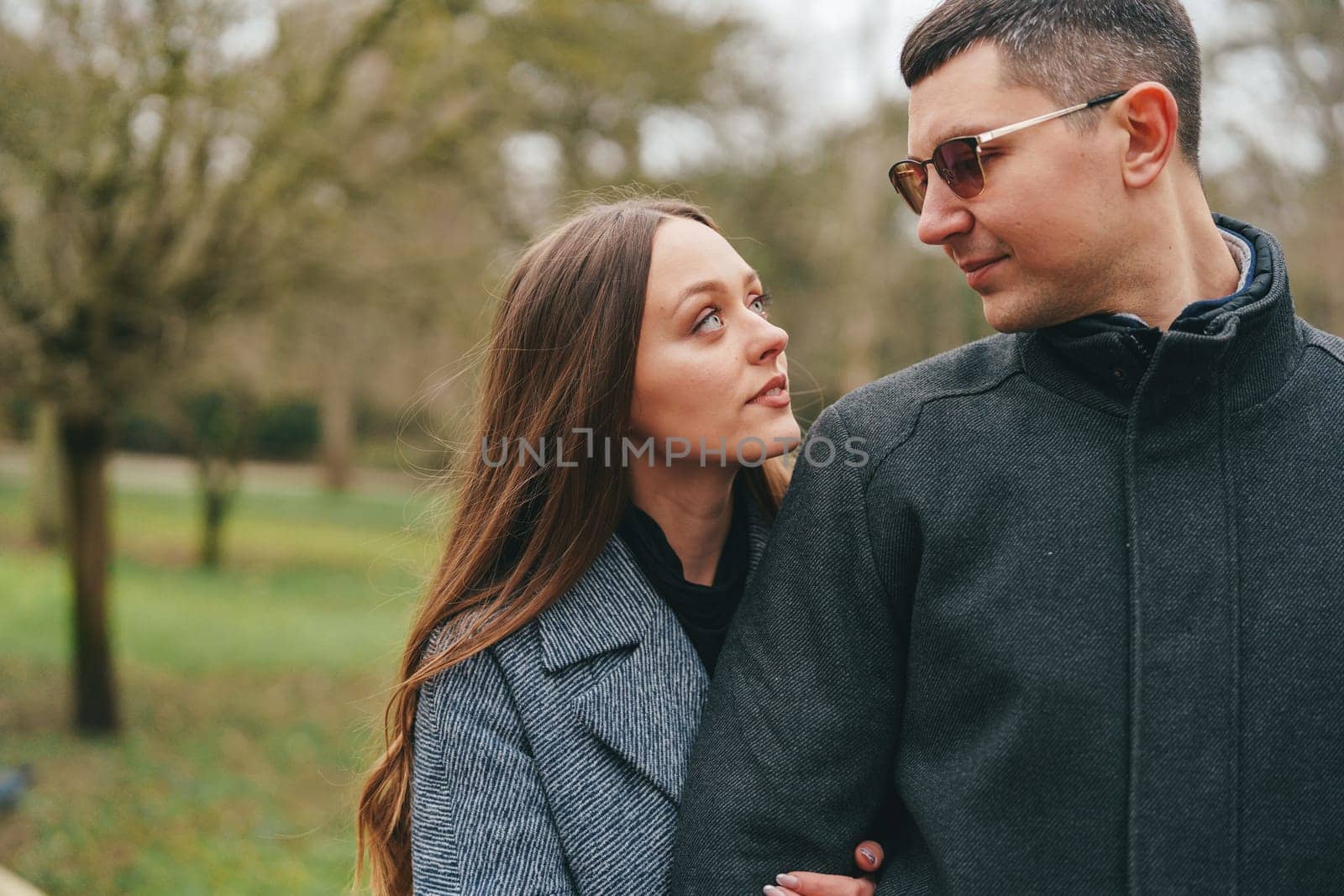 A couple stands close together in a lush park, sharing a tender moment. The woman gazes up at the man while he looks back warmly. Sunlight filters through the trees, creating a serene atmosphere.