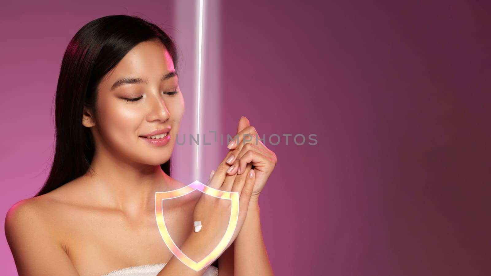 A woman applies a skincare product on her wrist, smiling as she enjoys the soothing experience. Soft pink lighting creates a calming atmosphere in the beauty studio.
