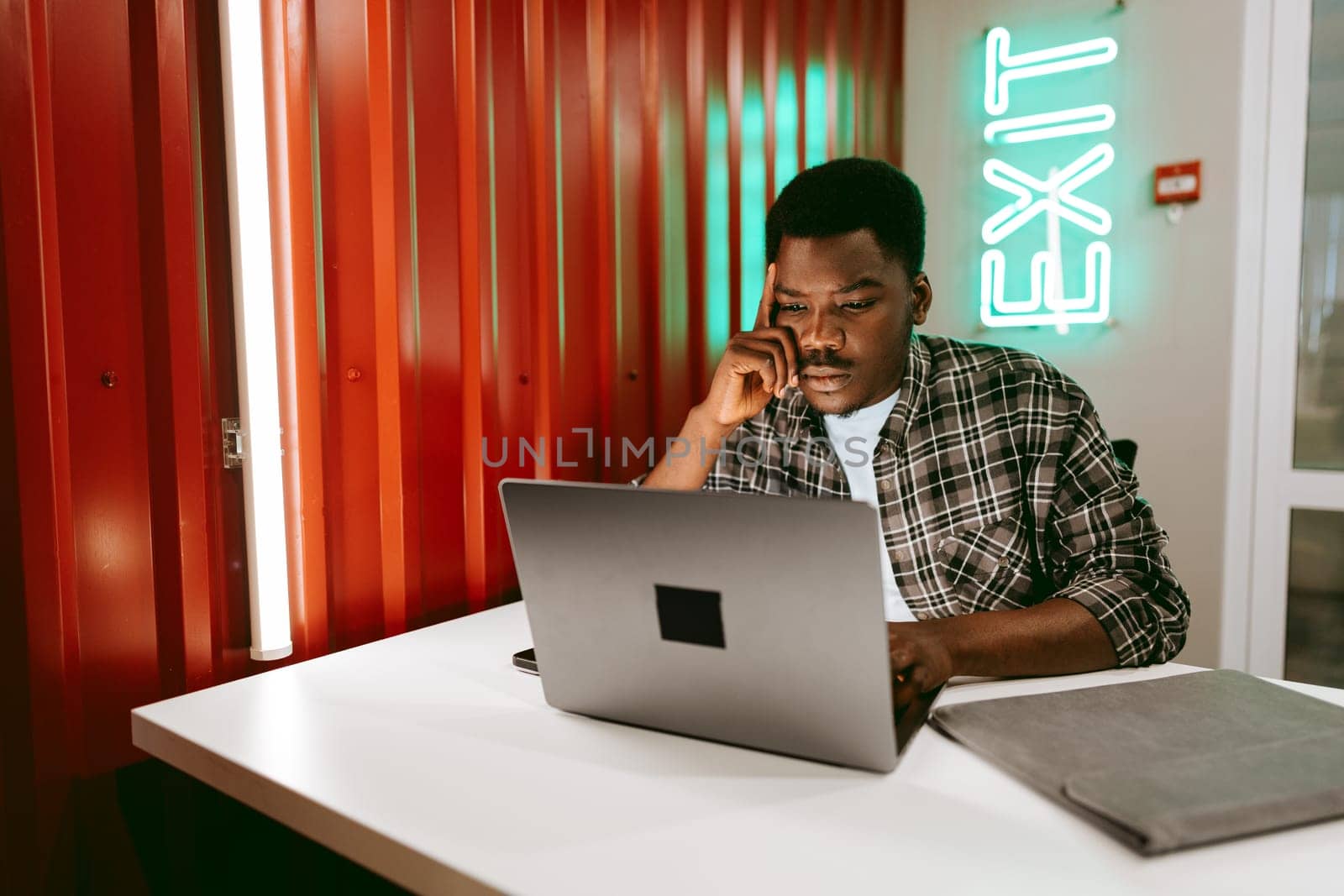 A young man wearing a checkered shirt is deeply focused on his laptop in a contemporary workspace. The vibrant neon exit sign adds a striking detail to the ambiance.