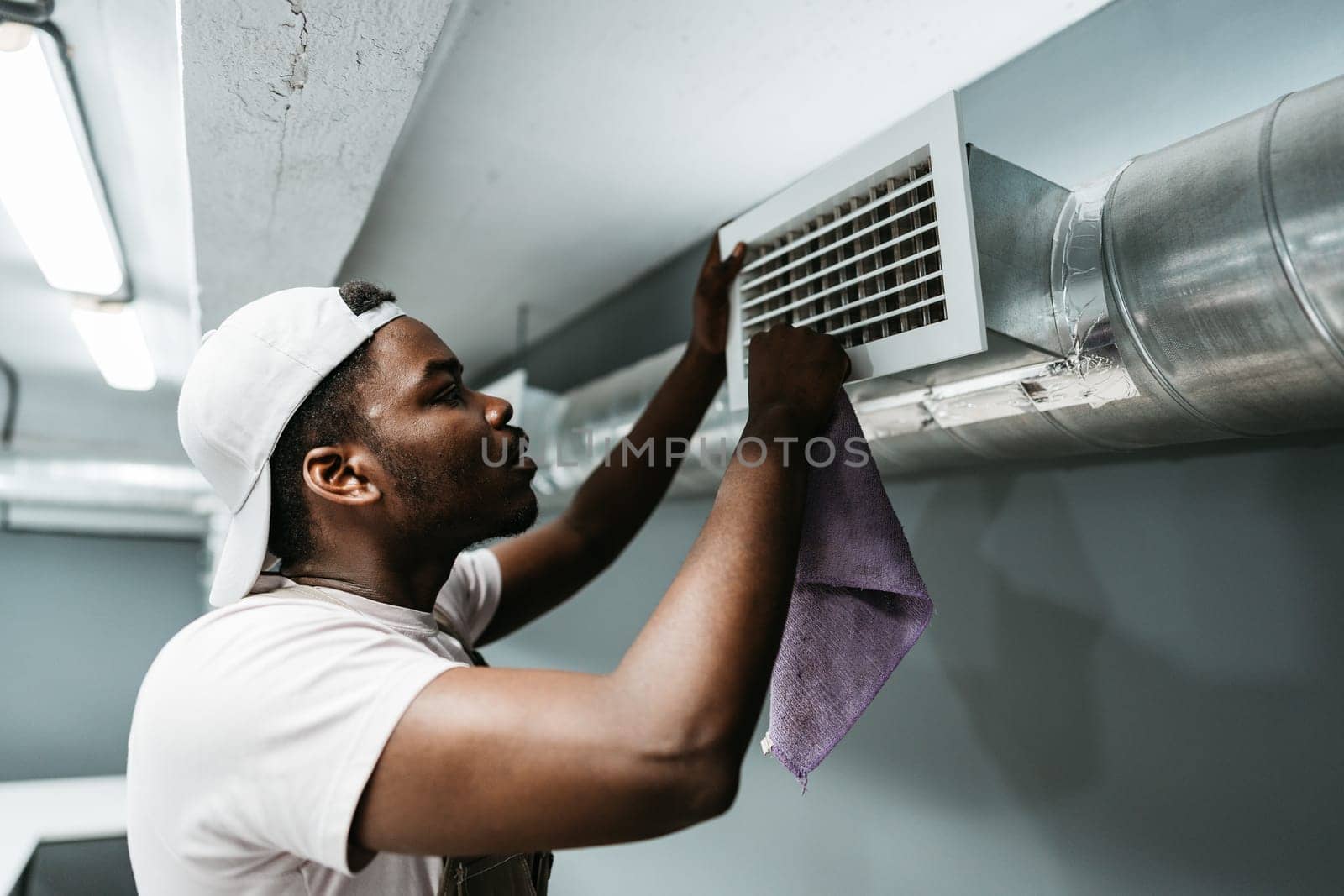 A man is focused on cleaning an air vent in a basement, using a cloth to remove dust and dirt. Proper maintenance ensures improved air quality in the enclosed space.