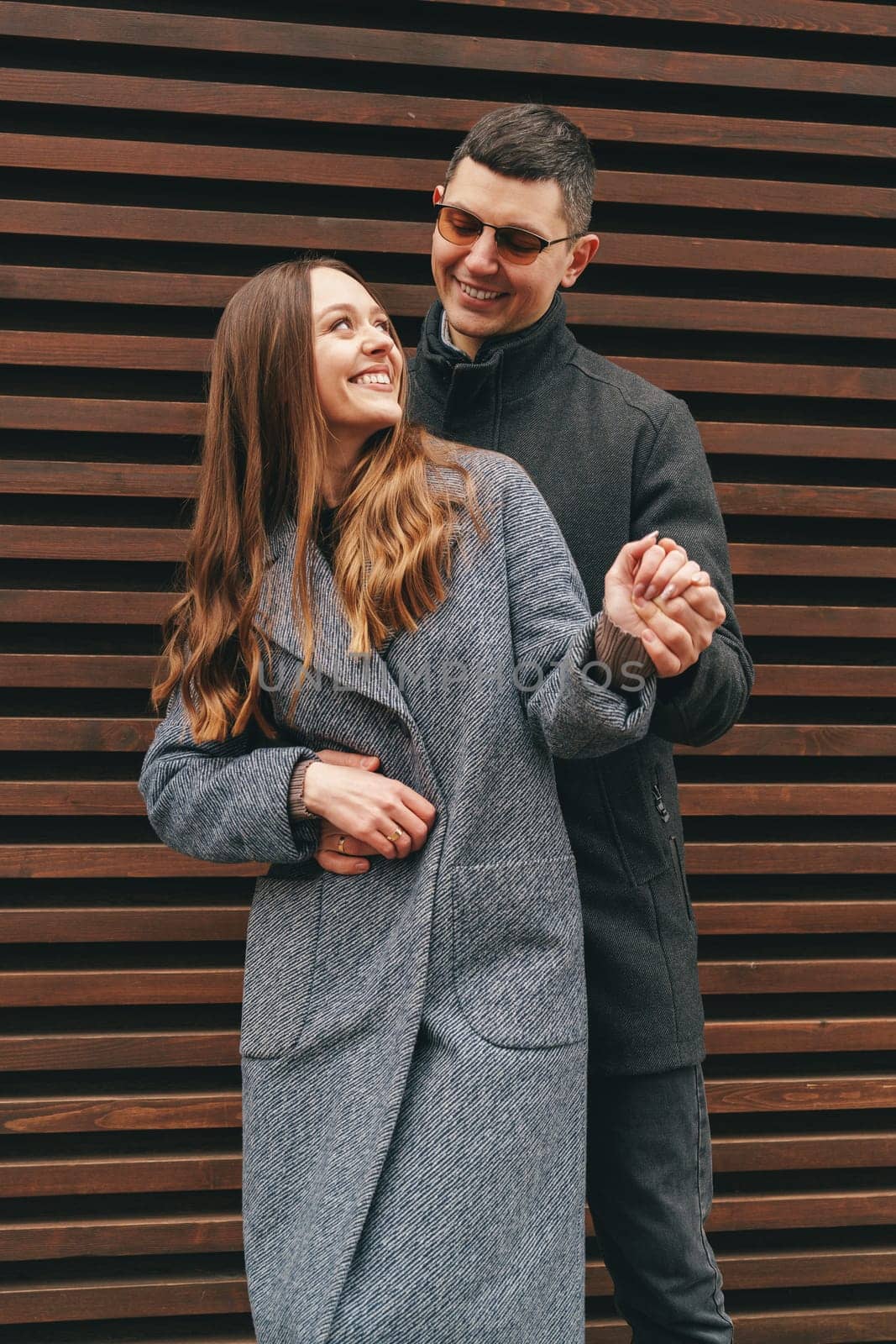 A couple smiles and embraces while standing in front of a textured wooden wall on a sunny day. The woman wears a fashionable coat, and the man is stylishly dressed.