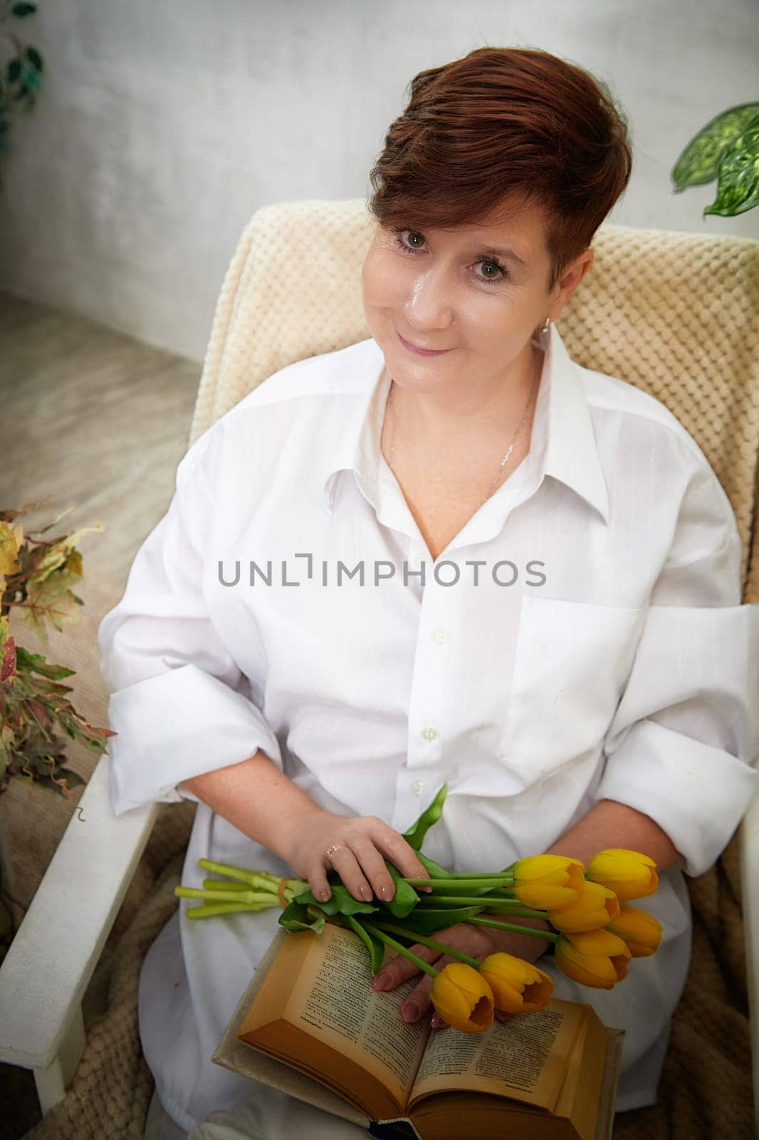 Middle-aged woman enjoys a relaxed moment with fresh tulips while reading a book in a cozy room on Women's Day.