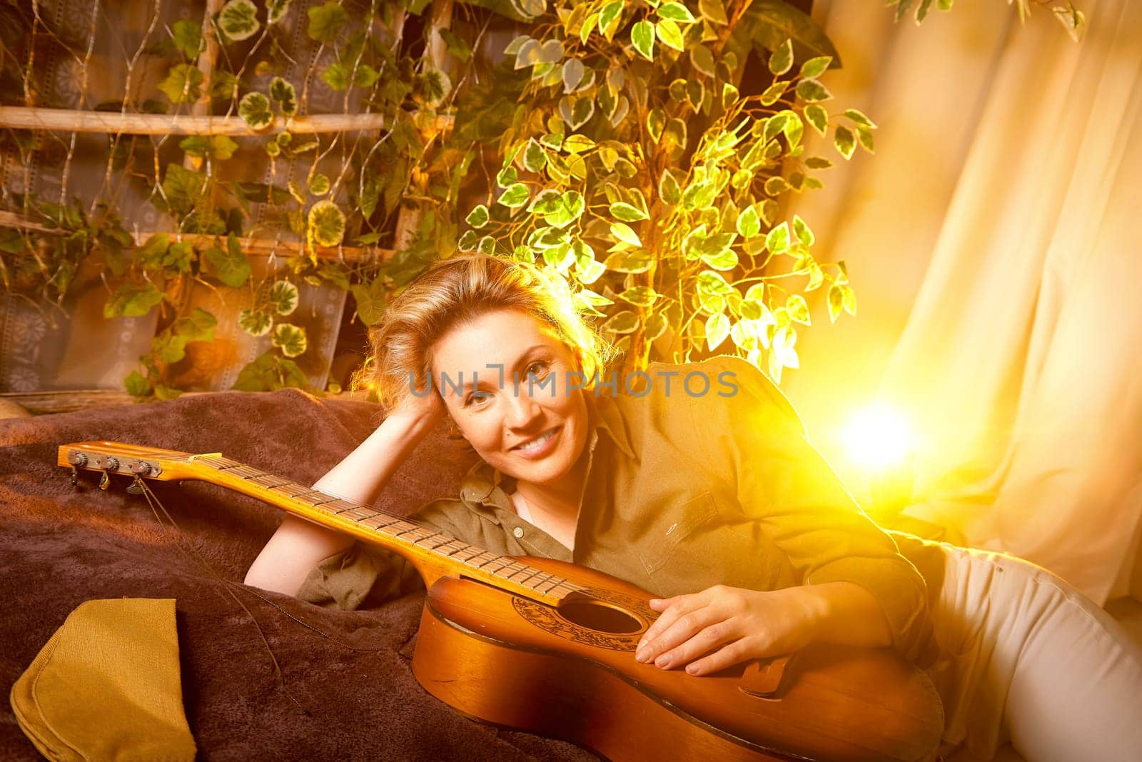 A musician lounges comfortably with their guitar beside glowing warm light and lush plants, creating a cozy atmosphere in a home setting.