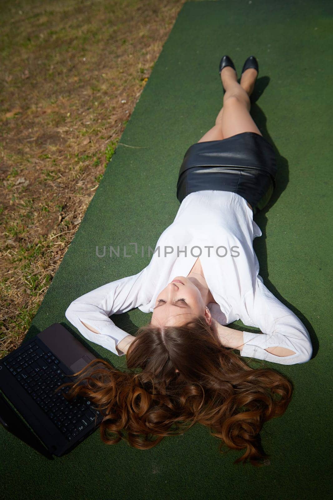 Beautiful woman in a white blouse and black skirt lies on green grass, using a laptop under bright sunlight.