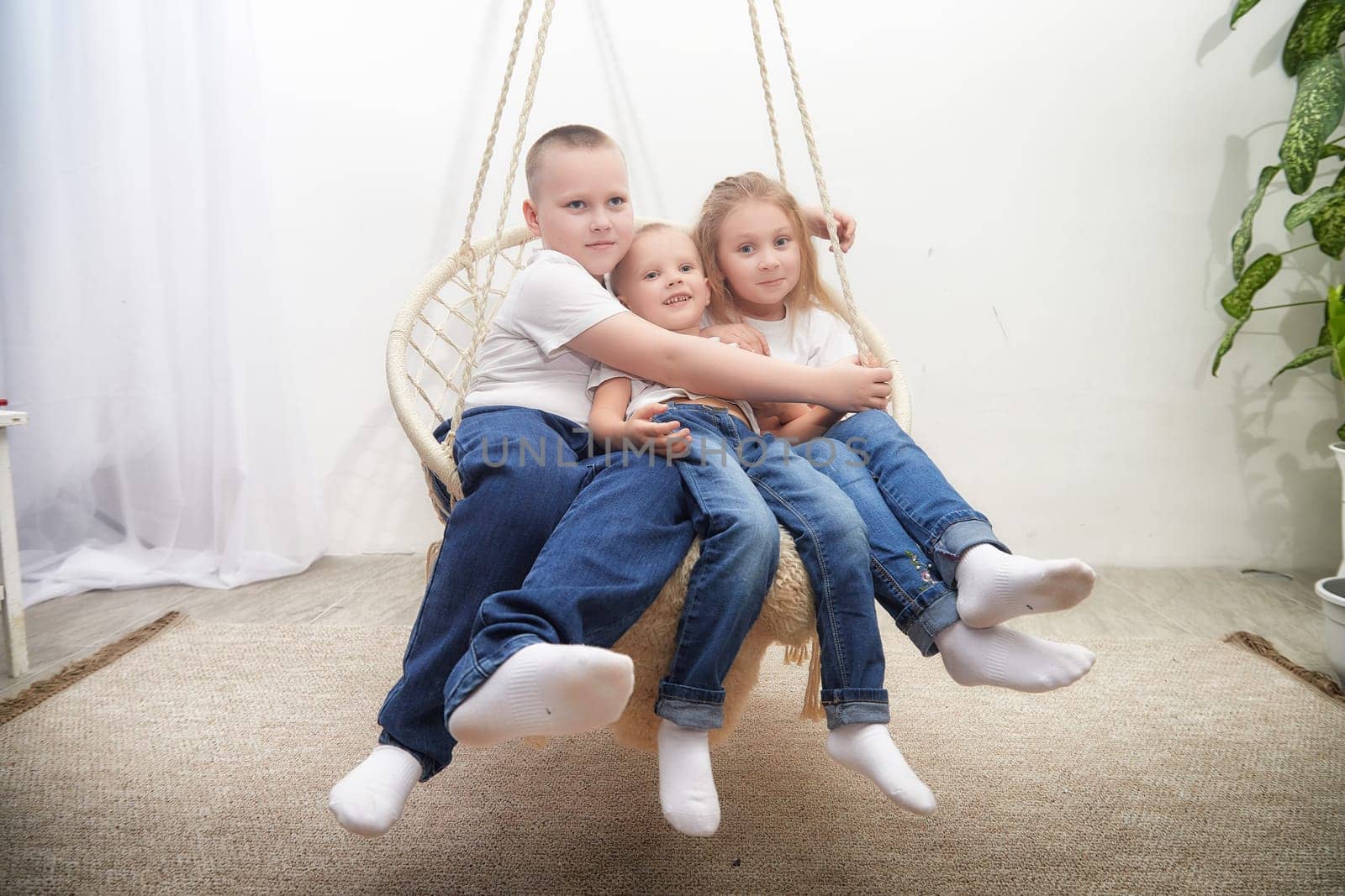 Three children sit closely together on a swing, smiling in a well-lit indoor space decorated with green plants.