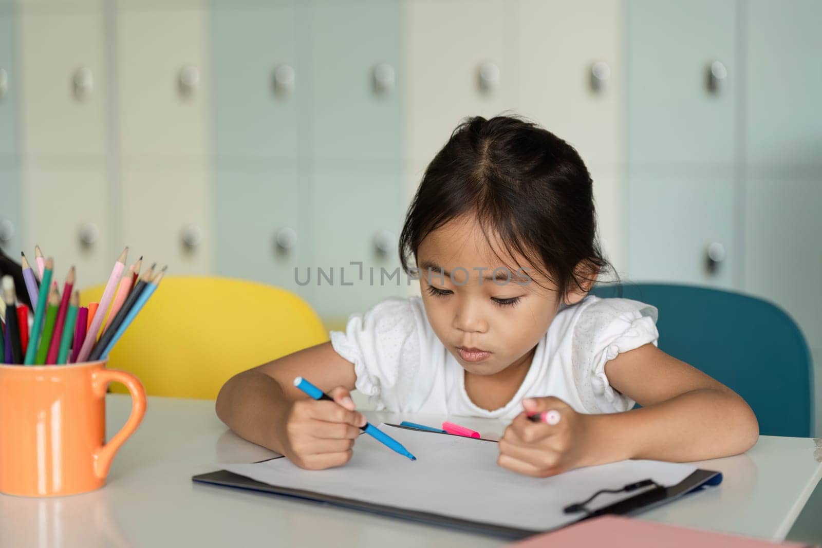 A young girl is deeply engaged in her art project, using vibrant colored pencils in a classroom setting. The atmosphere is creative and inspiring.