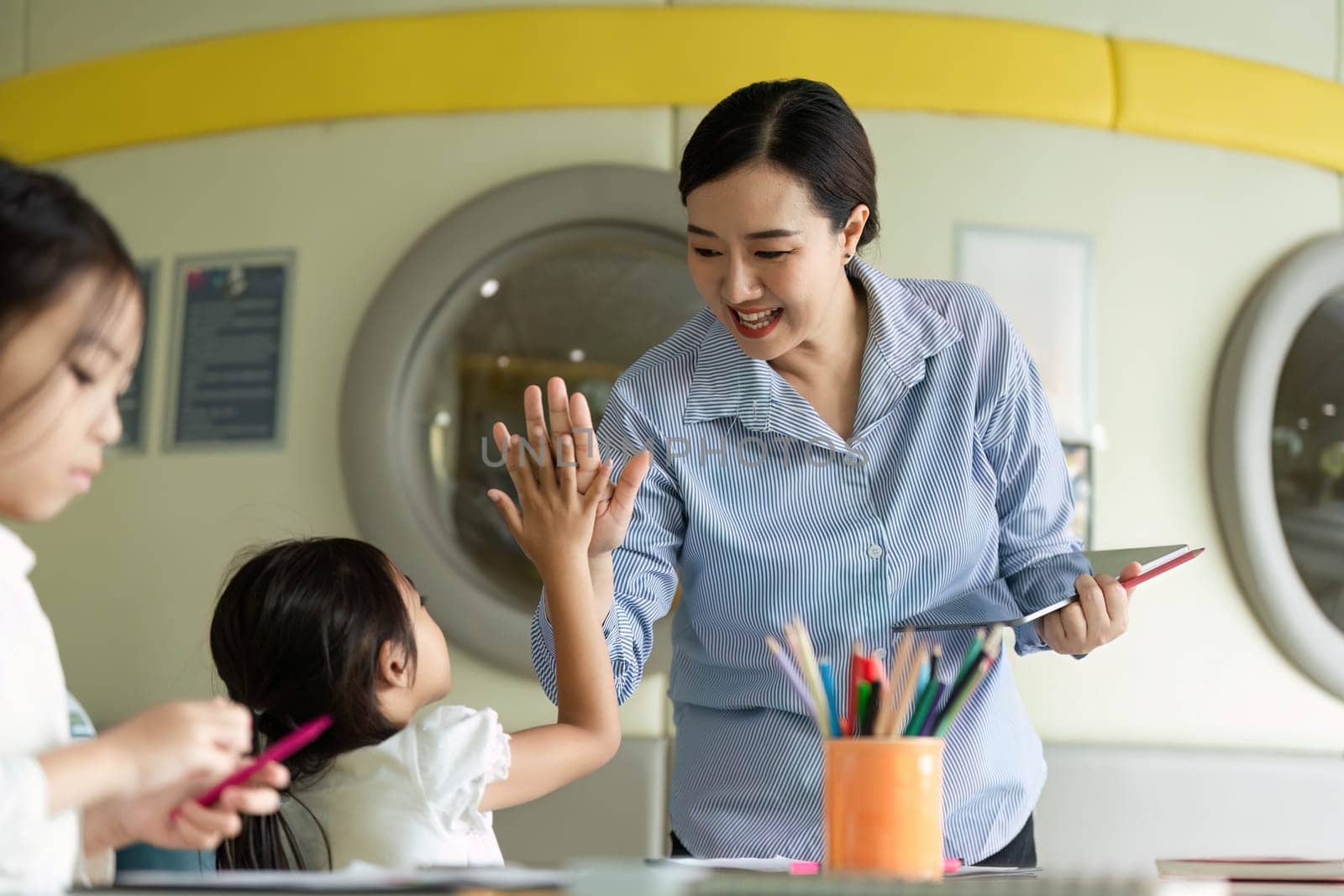 A teacher joyfully high-fives a young girl, celebrating her achievement in a colorful classroom filled with creativity and learning.