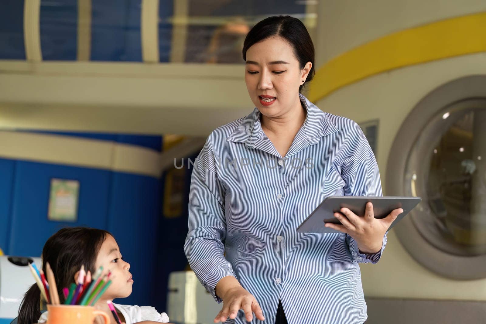 A teacher is actively engaging with her students, using a tablet to enhance learning in a colorful classroom environment.