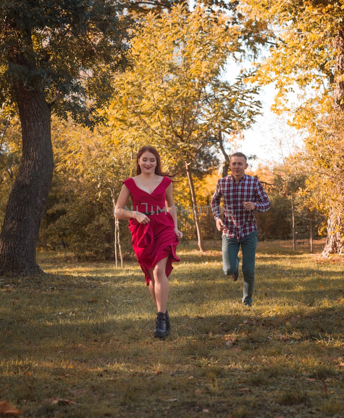 young couple in love actively relaxing in autumn park. they are running, having fun and laughing. girl in red dress, guy in checkered shirt against golden autumn background. concept of romance