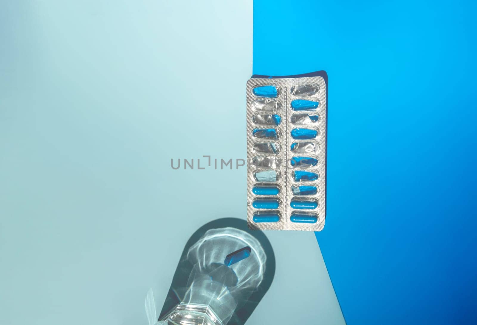 A clear glass of water casting a shadow on a blue surface and a blister pack of blue capsules, with several blue capsules placed nearby. Strong shadow and reflective effect through the glass