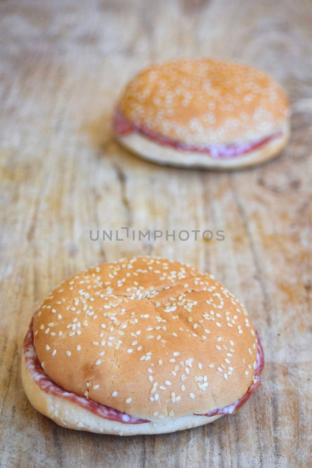 Close-up: hands using tongs to place salami slices on a bread roll. Another assembled salami sandwich in background on a wooden cutting board.