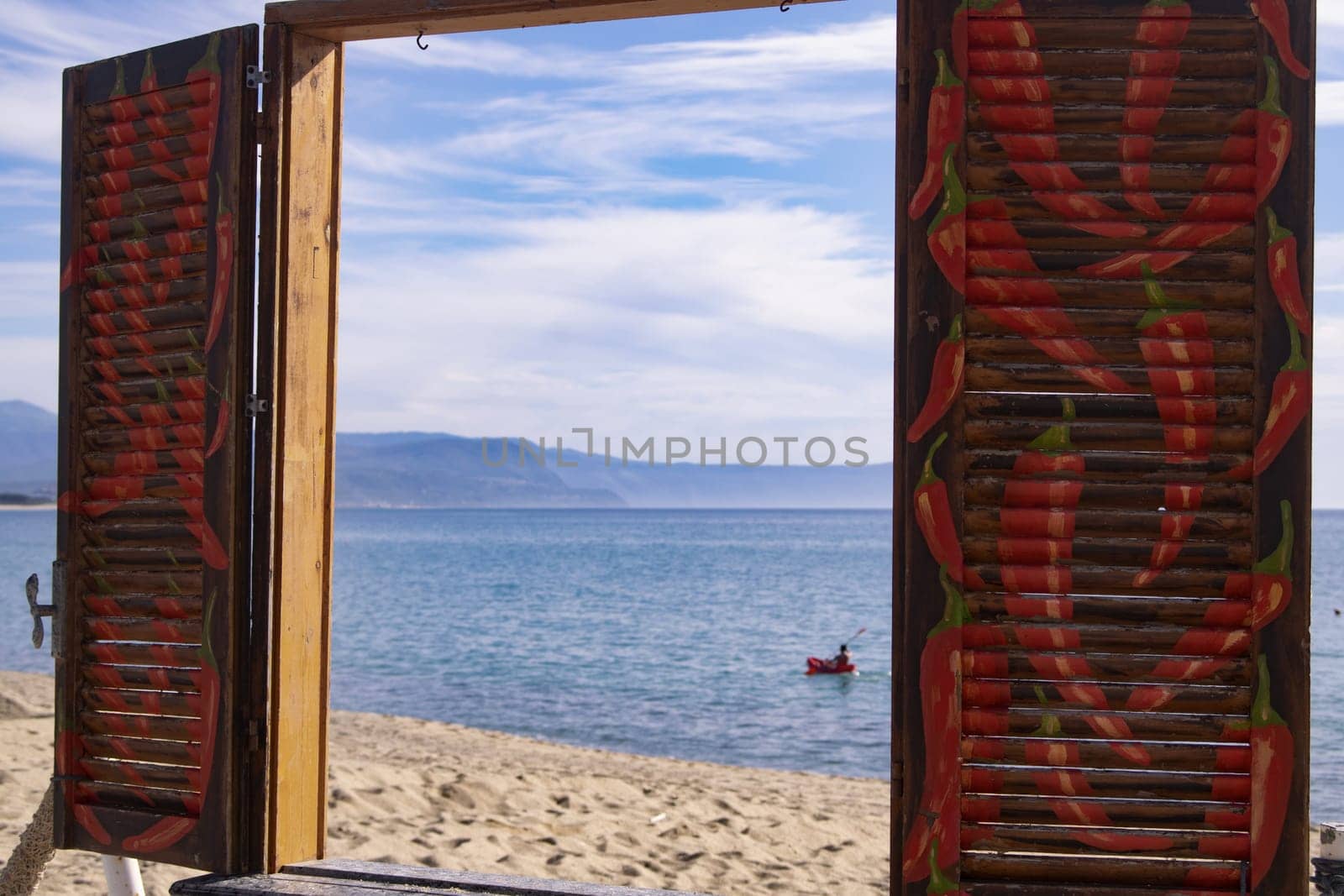 Vibrant blue sea & sandy beach framed by open, rustic wooden shutters with red chili peppers. Small boat in distance on serene water under partly cloudy sky.