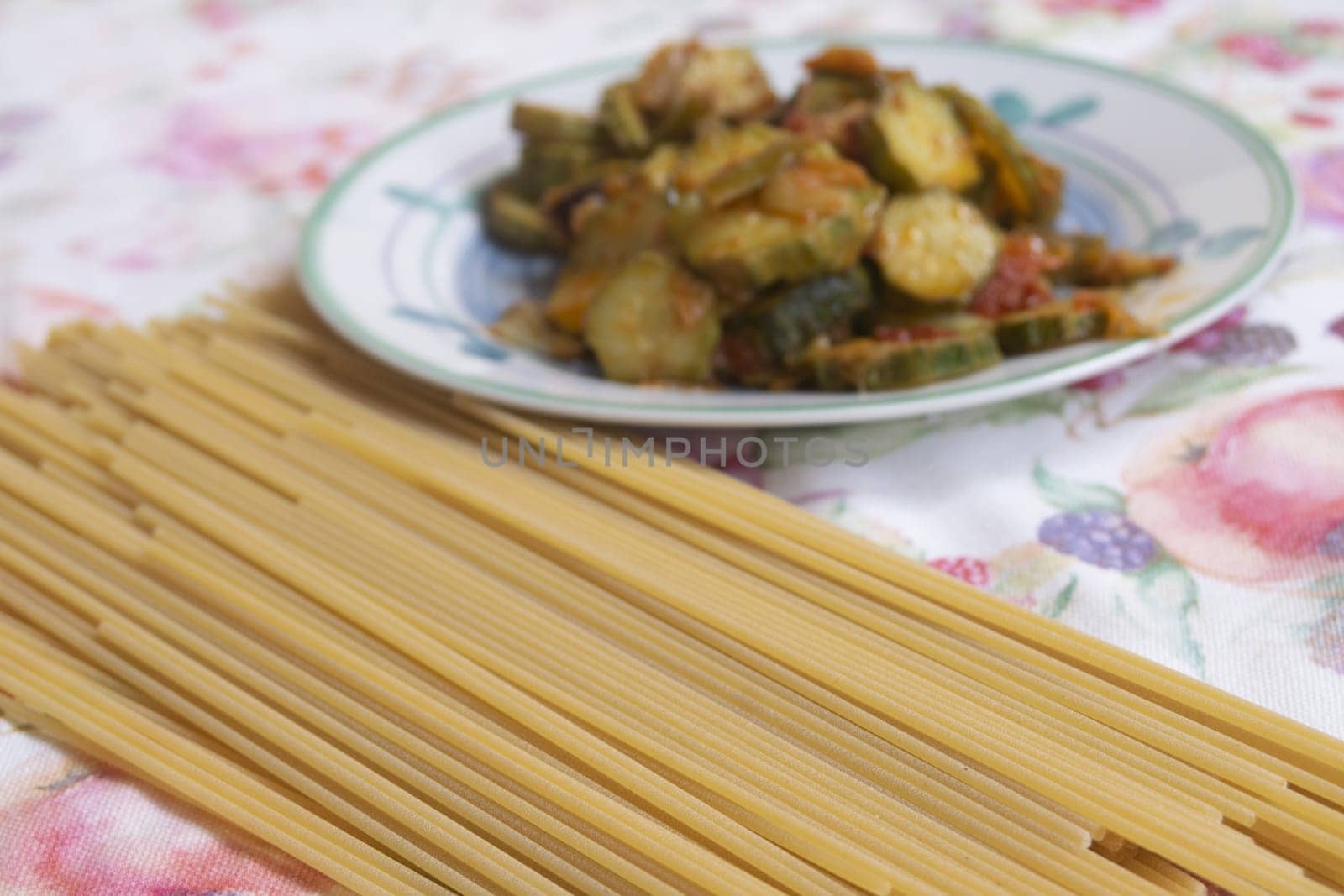 Uncooked whole wheat spaghetti on a floral tablecloth. Blurred background: notepad & cooked vegetables, suggesting healthy meal prep. Best for nutrition themes.