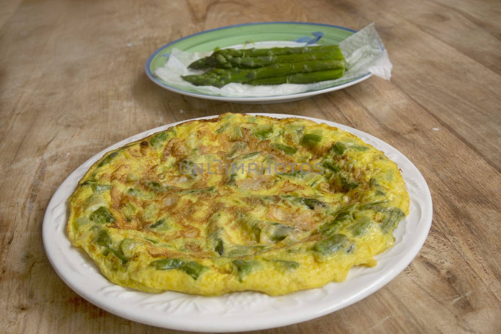 Homemade omelette with green asparagus & Parmesan, on a white plate on a rustic wooden table. Blanched asparagus in background. Perfect for healthy breakfast/brunch.