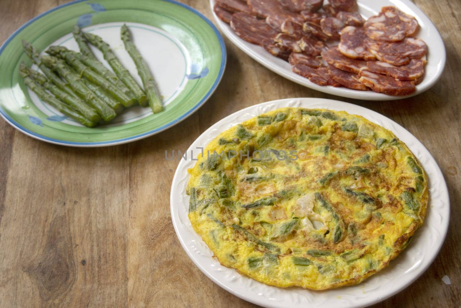 Golden omelette with fresh green asparagus on a white plate on a rustic wooden table. Blanched asparagus visible in background