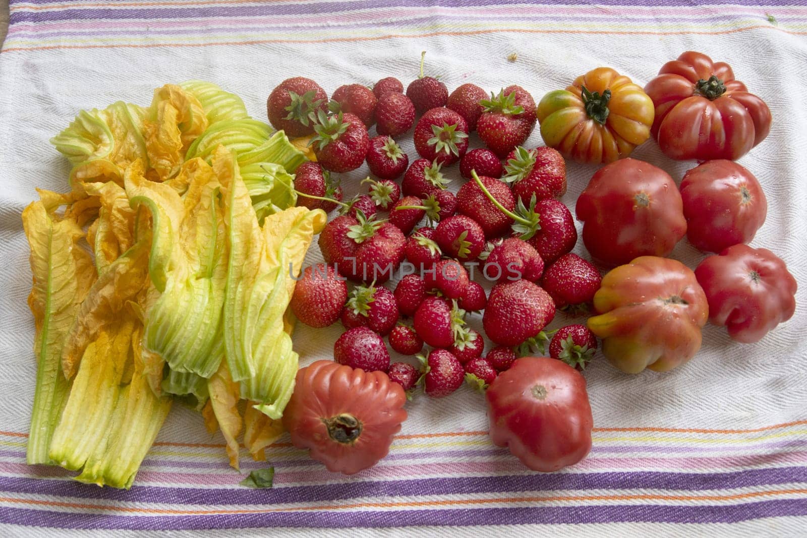 Vibrant summer bounty: ripe heirloom tomatoes, fresh strawberries, and delicate zucchini blossoms artfully arranged on rustic linen. Ideal for healthy eating, organic food, and farm-to-table themes