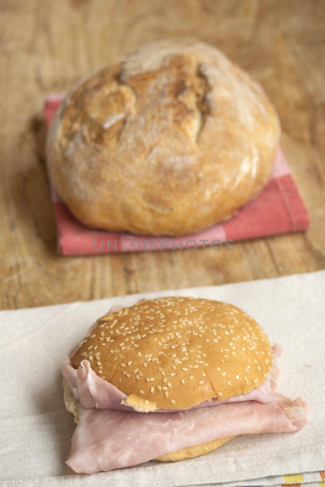 Classic ham sandwich on a sesame bun with a light napkin in foreground. Soft-focus background: rustic bread on checkered napkin. All on wood, conveying simple, homemade meals