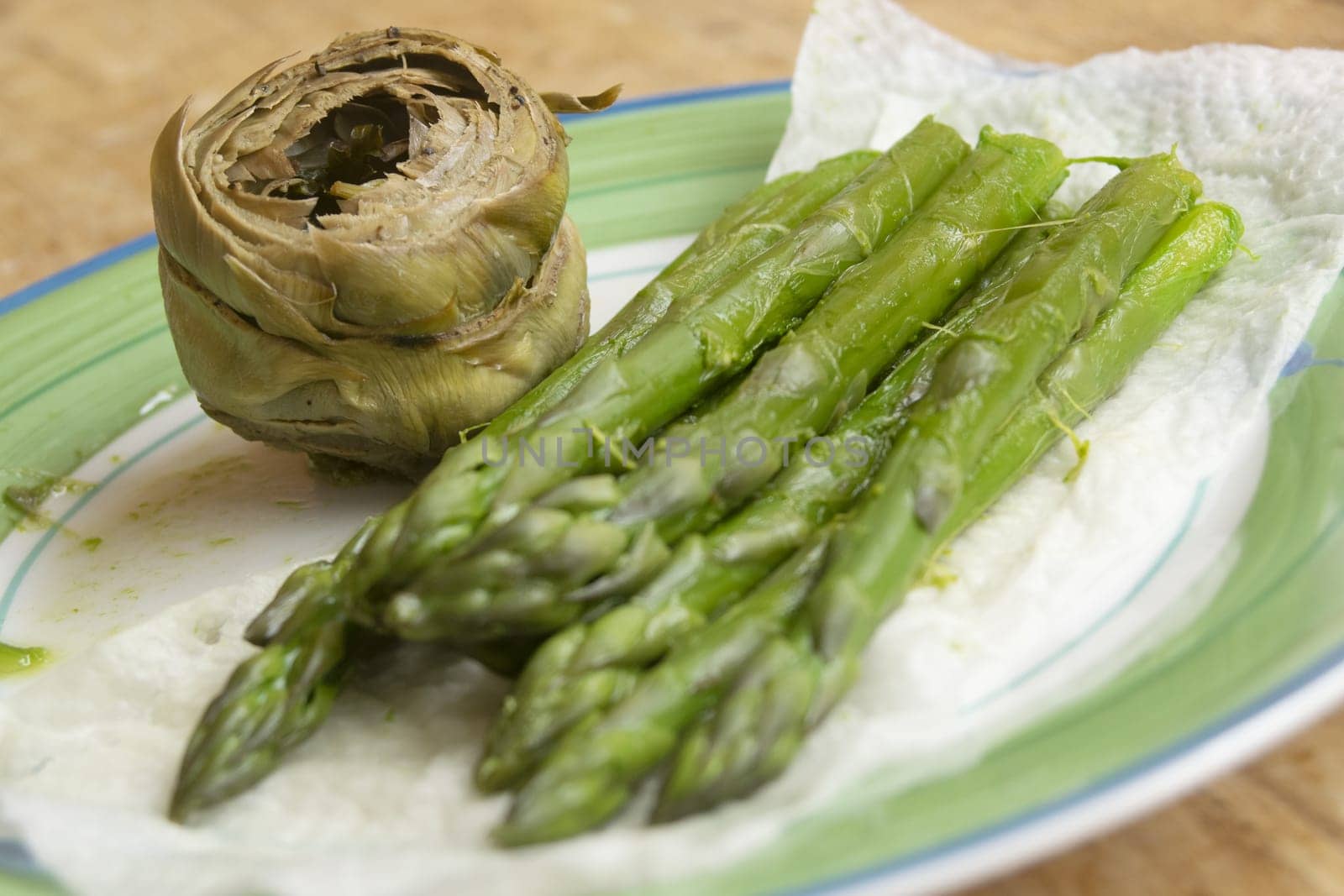  A close-up view of fresh, vibrant green asparagus spears resting on a paper towel, beside a cooked artichoke head on a light-colored plate. The focus is on the healthy, freshly prepared vegetables