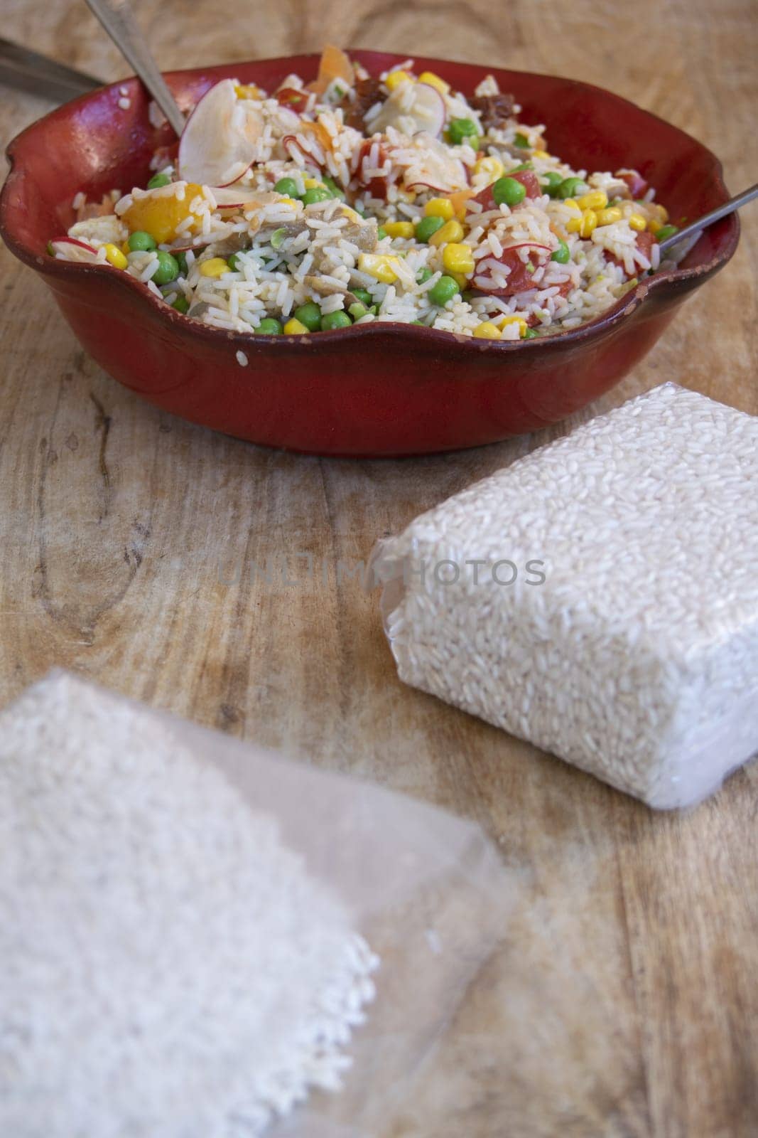 Colorful rice salad with fresh vegetables and uncooked rice on a wooden table, highlighting natural ingredients and healthy eating.