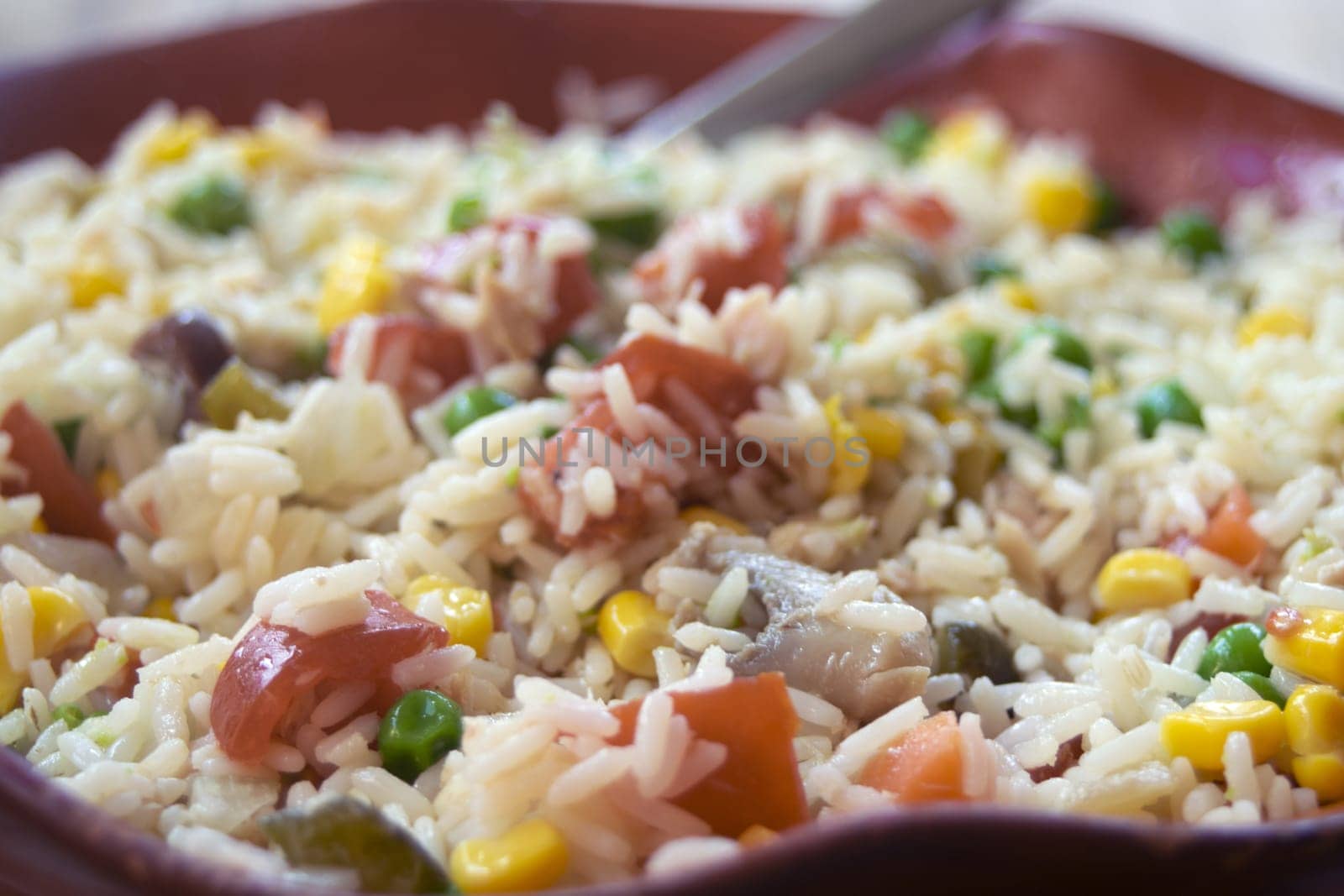 Close-up of a vibrant rice salad in a red bowl, featuring rice mixed with colorful vegetables like tomatoes, corn, peas, and possibly others. A spoon rests inside the bowl.

